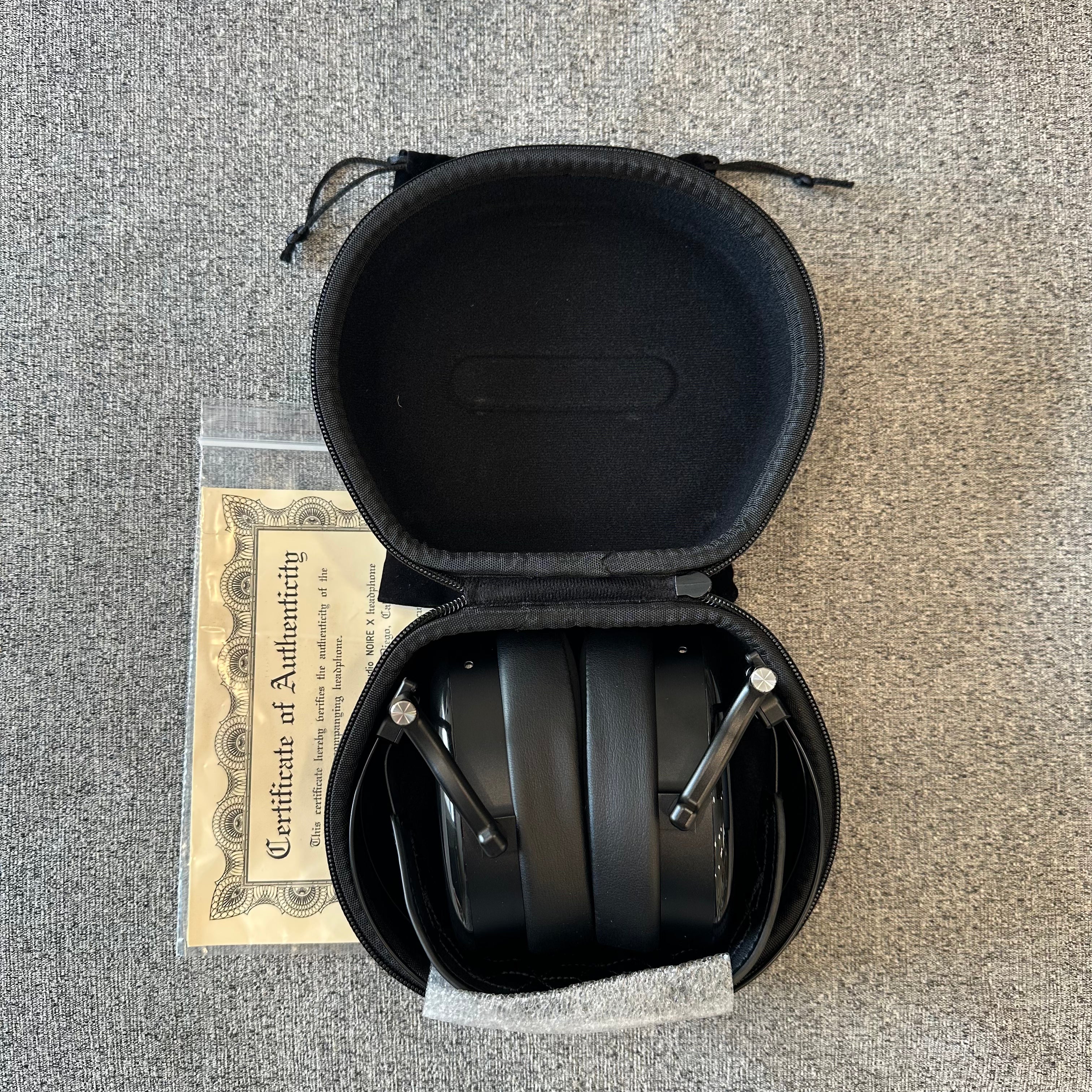Black headphones in an open case on a textured surface with a certificate of authenticity below.