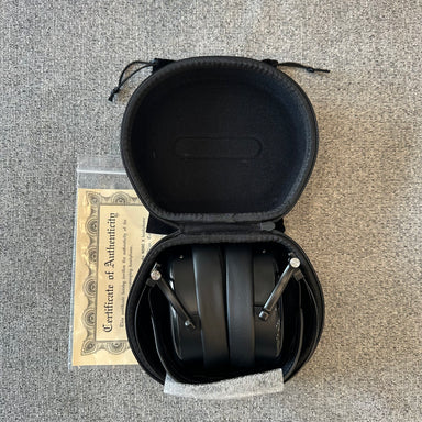 Black headphones in an open case on a textured surface with a certificate of authenticity below.