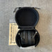Black headphones in an open case on a textured surface with a certificate of authenticity below.